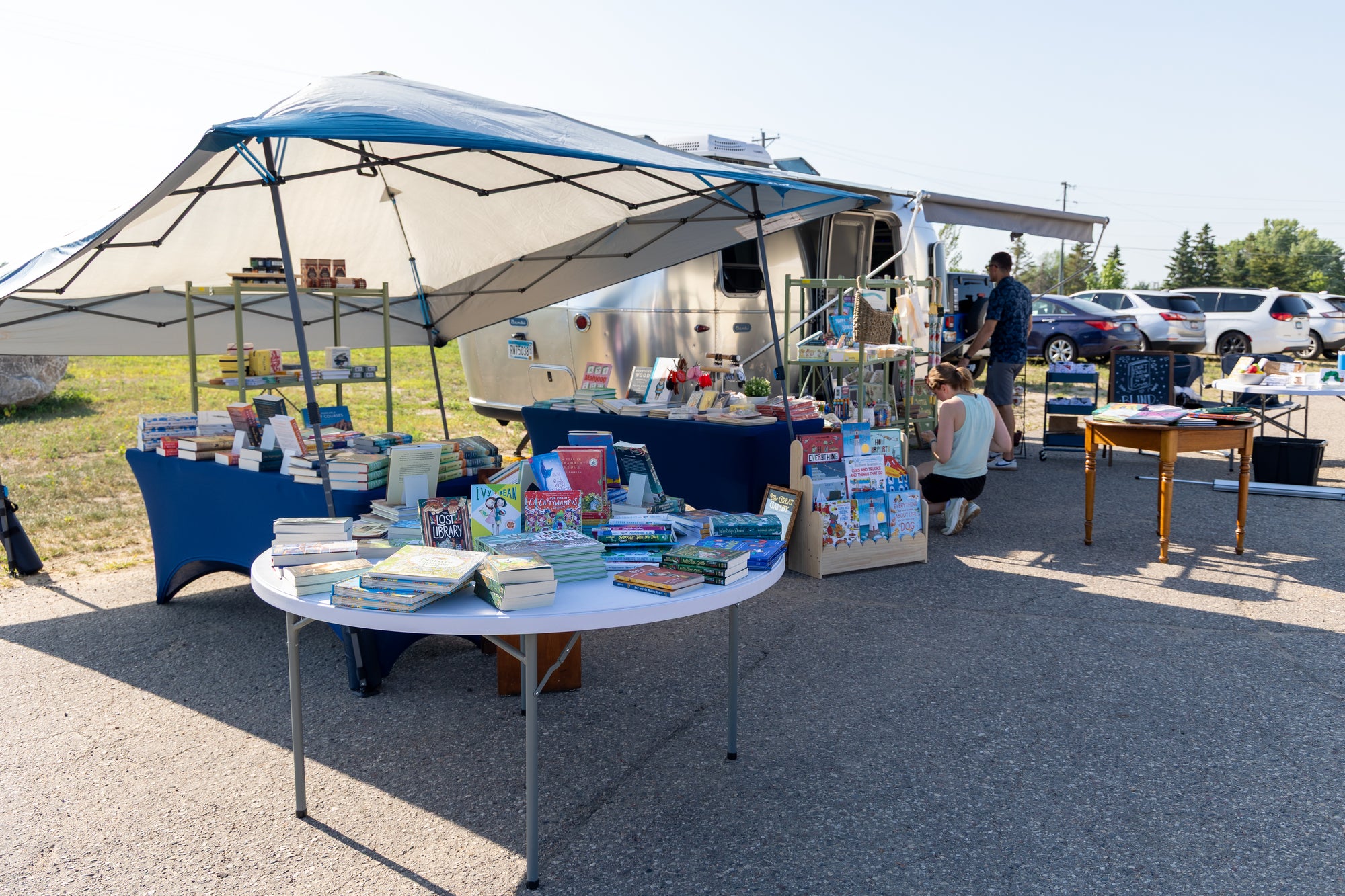 Outdoor market stall setup under a large white and blue canopy, with tables displaying books and other items in a paved lot. Several people are visible around the stall, and cars and trees appear in the background.